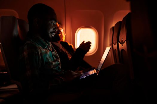 Male passenger browsing internet on laptop and flying abroad in airplane, using international airline to travel on business work trip. Young man working on computer and travelling during sunset.