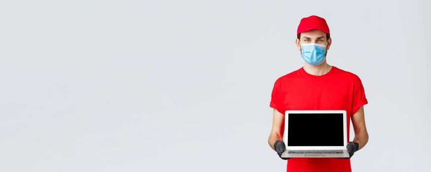 Customer support, covid-19 delivery packages, online orders processing concept. Smiling courier in red uniform, face mask and gloves, showing laptop screen webpage of company.