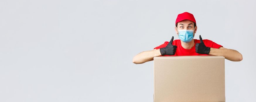 Packages and parcels delivery, covid-19 quarantine and transfer orders. Enthusiastic courier in red uniform, face mask and gloves, lean on box order and thumb-up, recommend service, express shipping.