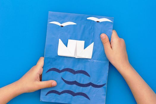 Flat lay child hand with paper craft greeting card with ship, sea and seagulls. Present for father s day