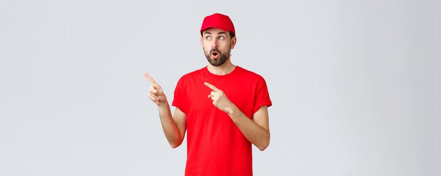 Online shopping, delivery during quarantine and takeaway concept. Impressed and wondered courier in red t-shirt with cap, employee uniform, open mouth shocked and staring left, pointing fingers.