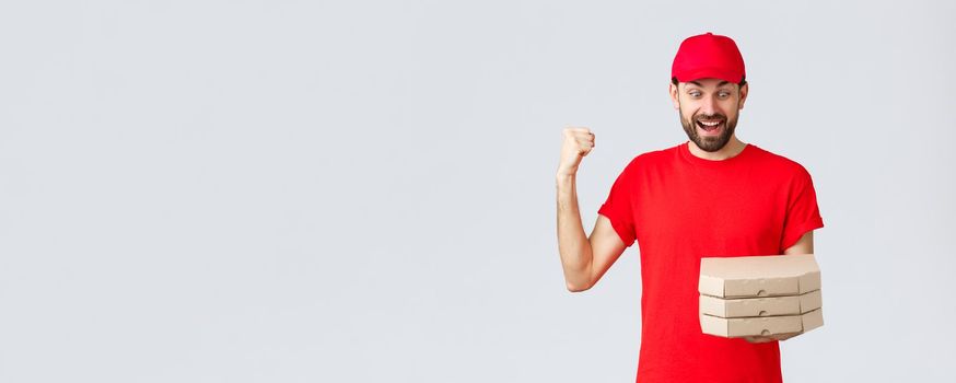 Food delivery, quarantine, stay home and order online concept. Excited and happy courier in red t-shirt and cap, rejoicing holding tasty pizzas, look at boxes and fist pump, grey background.