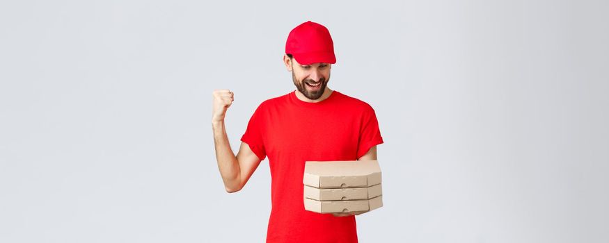 Food delivery, quarantine, stay home and order online concept. Happy, excited bearded courier in red t-shirt and cap celebrating, rejoicing, looking at pizza orders, grey background.