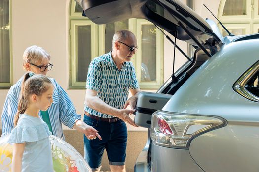 Retired people and niece loading baggage in trunk of automobile to leave on holiday road trip. Grandparents taking small child to seaside destination with luggage and inflatable during summer.