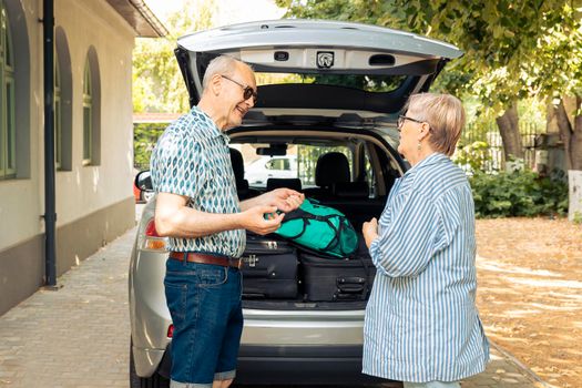 Old husband and wife sitting in driveway with car, leaving on holiday vacation adventure after loading travelling bags and suitcases in automobile trunk. Leaving on journey with trolley for leisure.