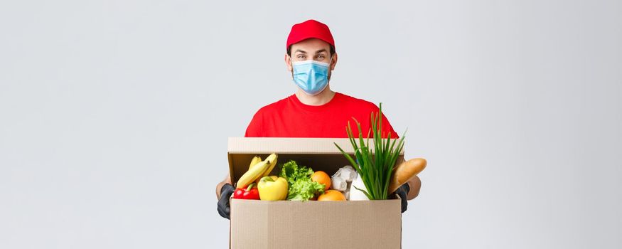 Groceries and packages delivery, covid-19, quarantine and shopping concept. Friendly courier in face mask and gloves delivering food box to client house during coronavirus, contactless deliver.