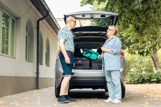 Elderly couple going on holiday vacation, loading suitcase and luggage in automobile trunk at home. Travelling by car with baggage and bags, leaving onubran cityscape adventure trip.