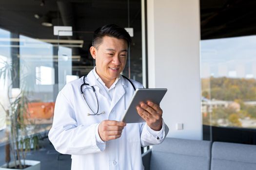 Asian doctor works in a modern clinic, in a private office, conducts an online consultation holding a tablet computer, reports good news, smiles at the camera, happy, video call