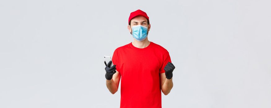 Contactless delivery, payment and online shopping during covid-19, self-quarantine. Rejoicing, happy courier in red uniform, gloves and mask, smiling, read good news in application, mobile phone.