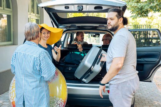 European family loading baggage in trunk of car to leave on seaside vacation with small kid, parents and grandparents. People prepaing luggage and travel bags to go on holiday journey.