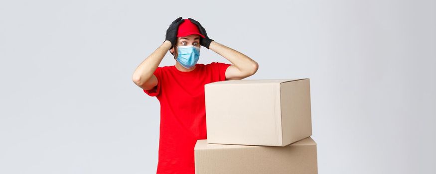 Packages and parcels delivery, covid-19 quarantine and transfer orders. Concerned and troubled courier in red uniform, face mask and gloves, grab head and gasping shocked staring at boxes.