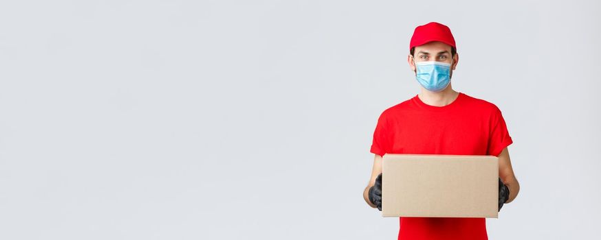Packages and parcels delivery, covid-19 self-quarantine delivery, transfer orders. Young courier in red uniform, gloves and face mask, holding box, give-out order to client, contactless service.