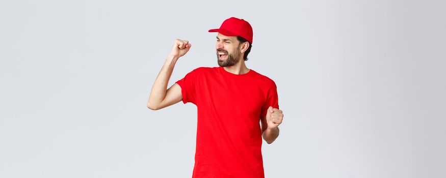 Cheerful and carefree young bearded delivery guy in red uniform cap and t-shirt, singing, yelling yes and fist pump in celebration, triumphing and rejoicing over great news, grey background.