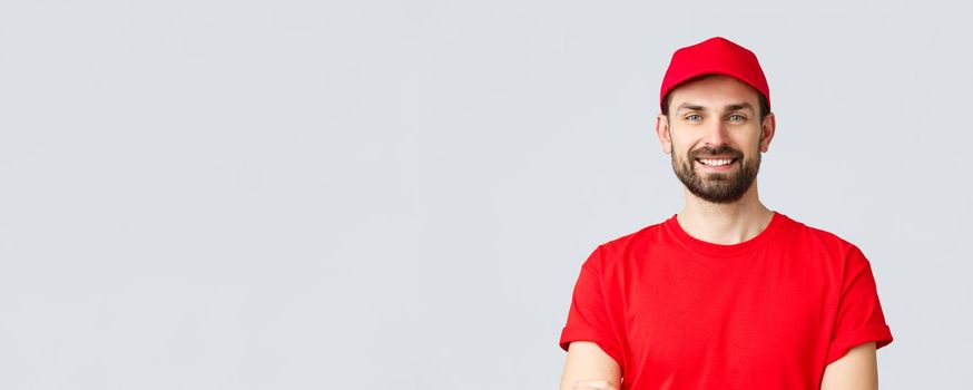 Online shopping, delivery during quarantine and takeaway concept. Handsome bearded courier in red uniform, smiling cheerful, cross arms chest, ready to deliver your order, grey background.