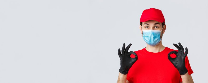 Contactless delivering, covid-19 and shopping concept. Cheerful, pleased courier in red uniform, cap and medical mask with gloves show okay, approval or guarantee gesture, recommend service.