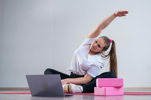Young overweight caucasian woman goes in for sports online by laptop. The girl sits on a mat and does side bends