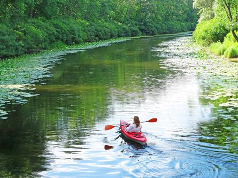 A girl dreamily floats her kayak down a river, surrounded by river plants. A romantic trip on the river alone. Thoughtful, atmospheric photo. Morning workout on the water