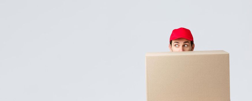 Packages and parcels delivery, covid-19 quarantine and transfer orders. Scared courier in red uniform cap, hiding behind customer order, looking left nervously, peeking at banner or advertisement.