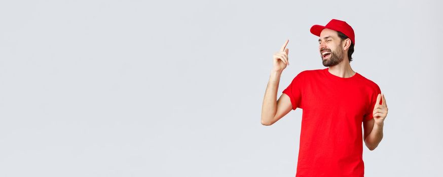 Good-looking carefree bearded man in red t-shirt and cap, uniform of employee, close eyes and dancing, pointing fingers up and singing. Delivery guy enjoying break at work, grey background.