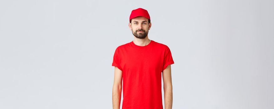 Online shopping, delivery during quarantine and takeaway concept. Young bearded courier in red uniform, employee wearing cap and t-shirt, looking at camera, waiting to take your order.