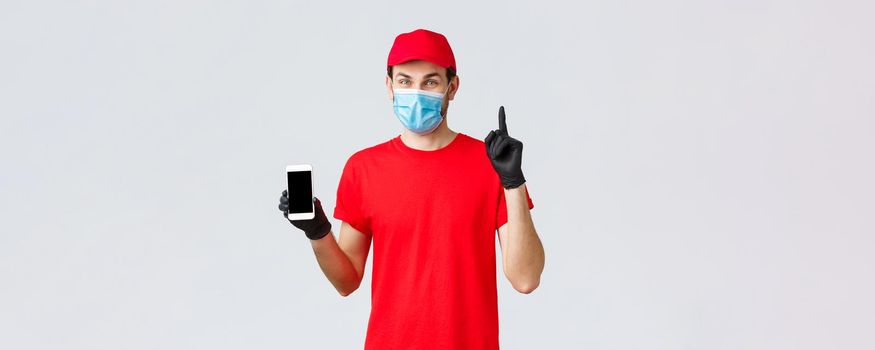 Contactless delivery, payment and online shopping during covid-19, self-quarantine. Friendly courier in red uniform, mask and gloves, pointing finger up, show smartphone display, order in internet.