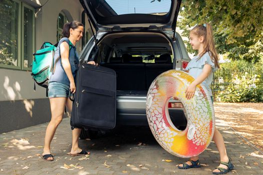 Mother and little girl leaving on vacation, loading luggage, bags and inflatable in trunk of automobile to travel at seaside. Family travelling on holiday journey with car during summer.