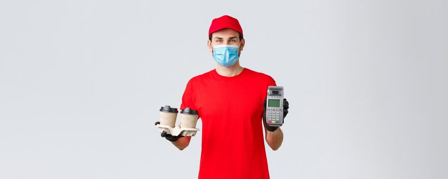 Takeaway, food and groceries delivery, covid-19 contactless orders concept. Friendly employee in red uniform, courier face mask and gloves, giving client POS terminal with coffee.