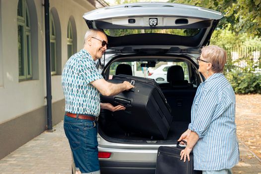 Old man and woman loading suitcase in car trunk, preparing to leave on road trip holiday. Travelling together during summer, going on vacation journey with luggage, baggage and bags.