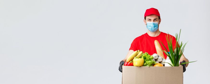 Groceries and packages delivery, covid-19, quarantine and shopping concept. Smiling courier in face mask, gloves and red uniform holding box with food, customer order, client home delivery.