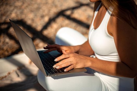 young beautiful woman with dark hair in white overalls sits on the sand near the sea and uses a laptop,