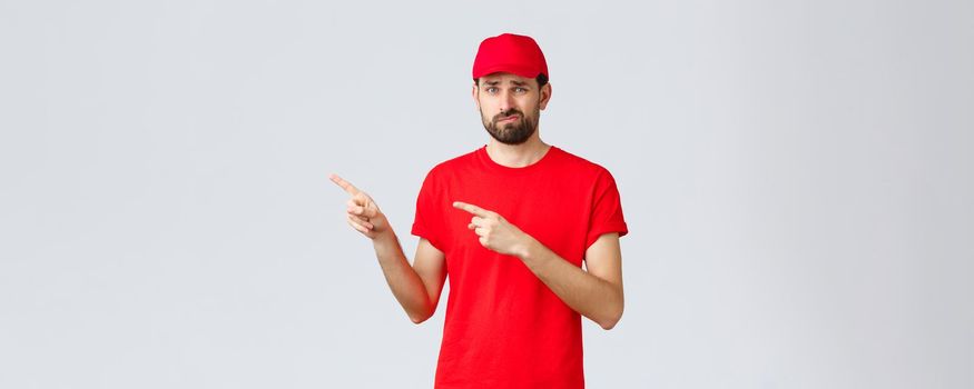 Online shopping, delivery during quarantine and takeaway concept. Shocked and impressed, gasping courier in red t-shirt and cap, uniform of service company, pointing fingers left astonished.