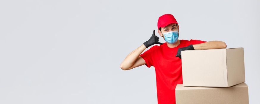 Packages and parcels delivery, covid-19 quarantine and transfer orders. Friendly courier in red uniform, face mask and gloves asking customer give call, show phone sign, stand near boxes.