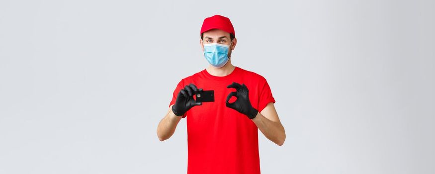 Contactless delivery, payment and online shopping during covid-19, self-quarantine. Friendly courier in red uniform, cap and t-shirt, wear face mask and gloves, thumb-up recommend credit card paying.