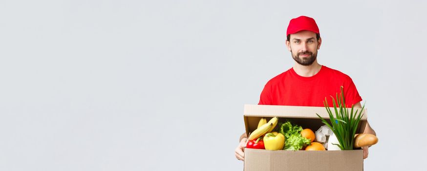 Groceries and packages delivery, covid-19, quarantine and shopping concept. Handsome courier in red uniform cap and t-shirt, bring grocery package, box delivery to client house, fast shipping.