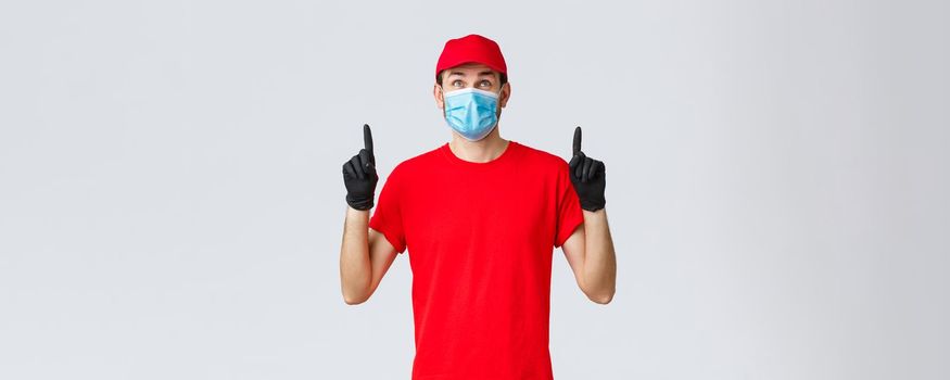 Covid-19, self-quarantine, online shopping and shipping concept. Smiling delivery guy red uniform cap and t-shirt, medical mask with rubber gloves, looking pointing up curious, reading sign or promo.