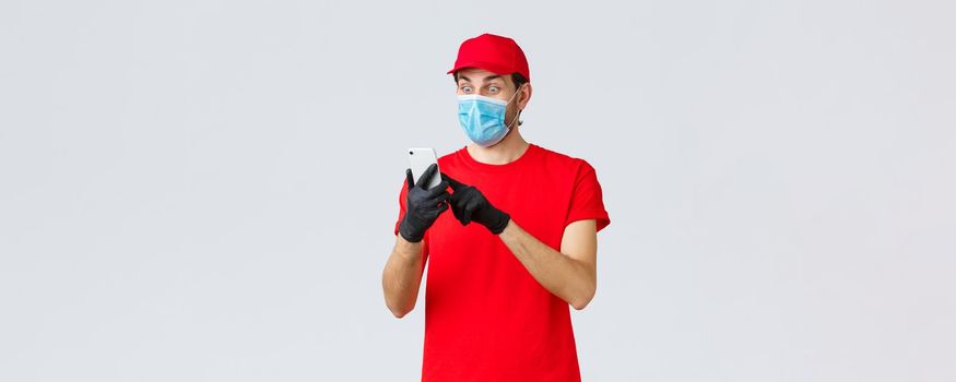 Contactless delivery, payment and online shopping during covid-19, self-quarantine. Excited courier in face mask and gloves, staring astonished at smartphone display, reading order info.