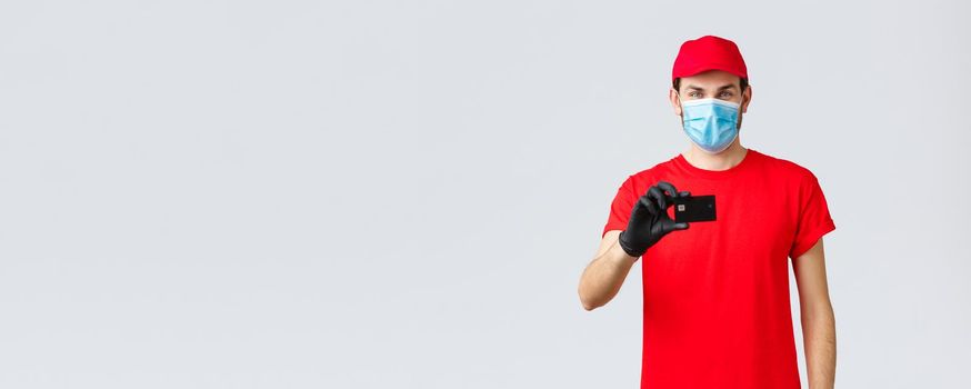 Contactless delivery, payment and online shopping during covid-19, self-quarantine. Handsome courier in red uniform, cap, medical face mask and gloves, show credit card, order internet.