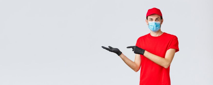 Groceries and packages delivery, covid-19, quarantine and shopping concept. Friendly courier in red uniform, face mask and gloves, introduce promo on banner, pointing left, showing advertisement.