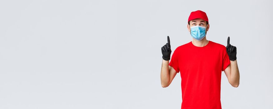 Covid-19, self-quarantine, online shopping and shipping concept. Smiling delivery guy red uniform cap and t-shirt, medical mask with rubber gloves, looking pointing up curious, reading sign or promo.