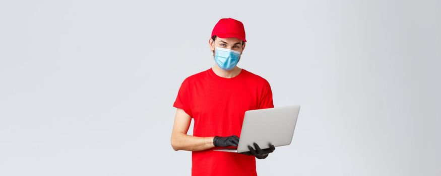 Customer support, covid-19 delivery packages, online orders processing concept. Pleasant delivery guy in red uniform, face mask and gloves, using laptop and look camera, processing order.