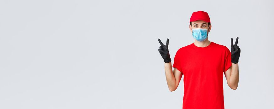 Covid-19, self-quarantine, online shopping and shipping concept. Cute delivery guy in red uniform, face mask and gloves, stay positive in coronvirus outbreak, show peace sign, courier deliver goods.