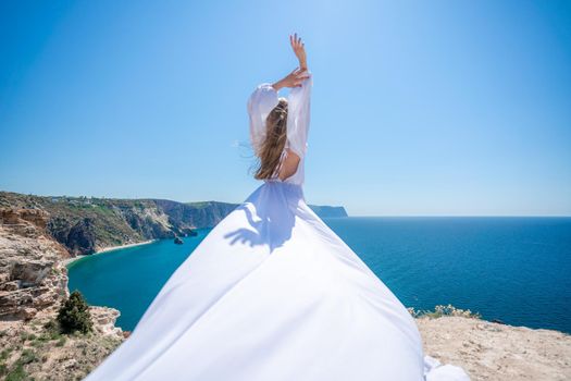 Blonde with long hair on a sunny seashore in a white flowing dress, rear view, silk fabric waving in the wind. Against the backdrop of the blue sky and mountains on the seashore