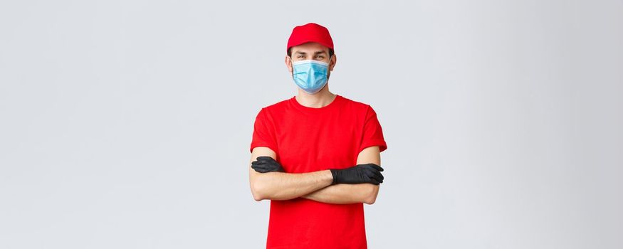 Covid-19, self-quarantine, online shopping and shipping concept. Confident smiling delivery man in red cap, t-shirt, wearing protective medical mask and rubber gloves while making courier order.