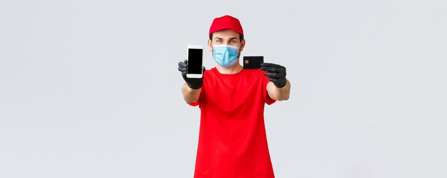 Contactless delivery, payment and online shopping during covid-19, self-quarantine. Friendly courier in red uniform, face mask and gloves, showing smartphone screen and credit card, order internet.