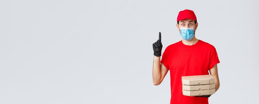 Food delivery, application, online grocery, contactless shopping and covid-19 concept. Excited delivery guy in red uniform, gloves and face mask, have suggestion, hold pizza and raise finger.