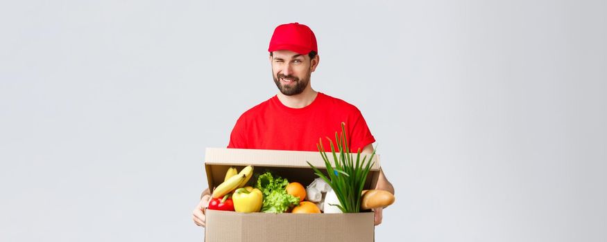 Groceries and packages delivery, covid-19, quarantine and shopping concept. Handsome smiling courier in red uniform, give cheeky wink as delivering food box, online order to client house.