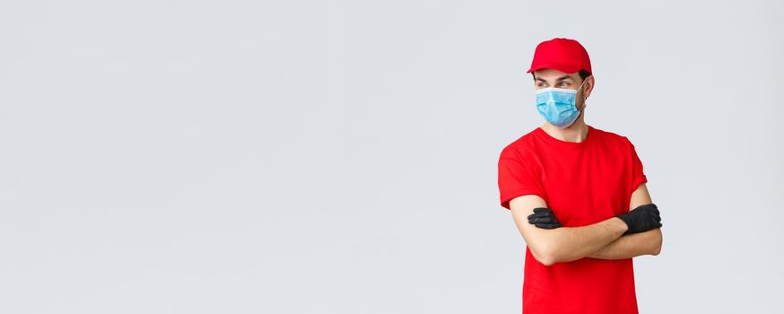 Covid-19, self-quarantine, online shopping and shipping concept. Confident courier in red uniform, protective gloves and face mask, standing crossed arms, look away, ready take your orders.