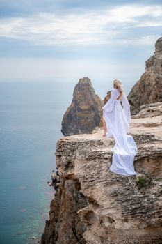 Blonde with long hair on a sunny seashore in a white flowing dress, rear view, silk fabric waving in the wind. Against the backdrop of the blue sky and mountains on the seashore