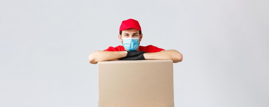 Packages and parcels delivery, covid-19 quarantine and transfer orders. Young courier in red uniform cap, face mask and gloves, leaning on box to deliver, shipping your orders, grey background.