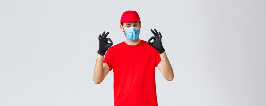 Covid-19, self-quarantine, online shopping and shipping concept. Handsome delivery guy in red cap, t-shirt, wear protective face mask and gloves from coronavirus, show okay, agree and good gesture.